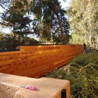 Wooden fence stained a warm brown, with trees and greenery in the background. A person stands nearby.