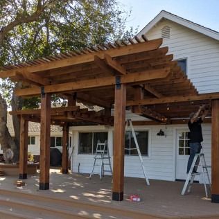 Wooden pergola attached to a white house with a man on a ladder installing lights.