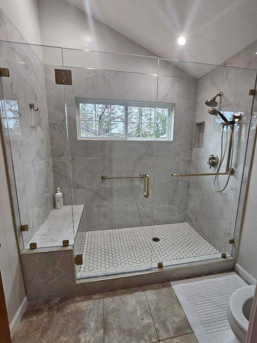 Glass shower with marble walls, gold fixtures, bench, and pebble floor.
