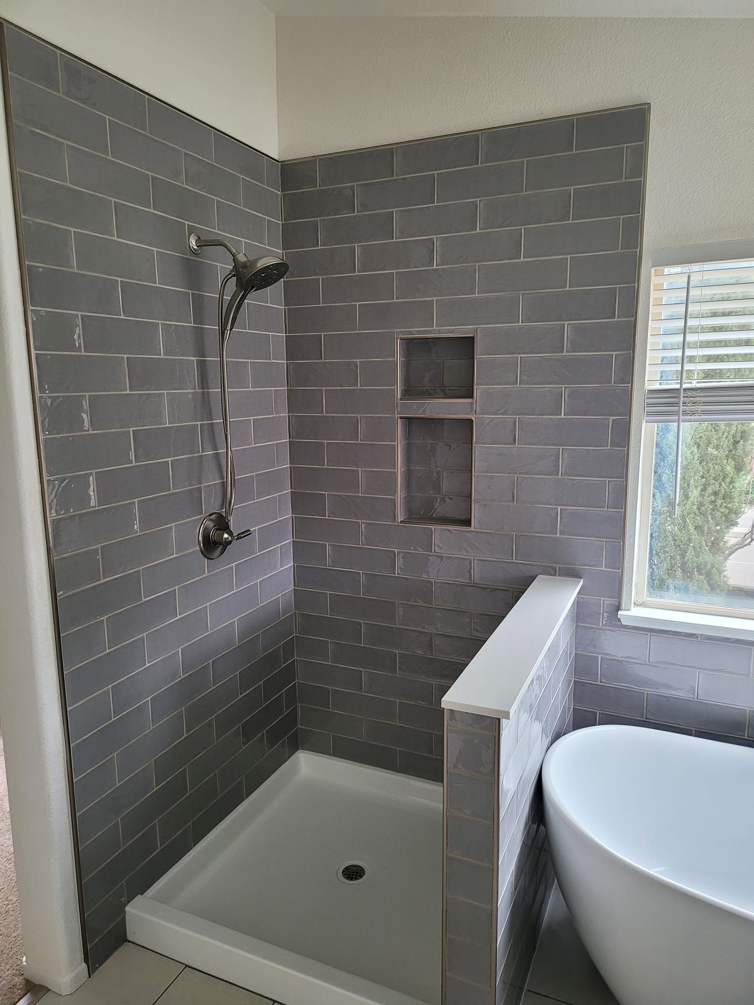 Grey tiled shower with a built-in shelf, showerhead, and white base, next to a bathtub and window.