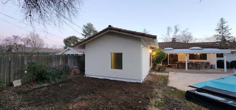Backyard with small white shed, wooden fence, patio with table, and pool. Overcast sky.