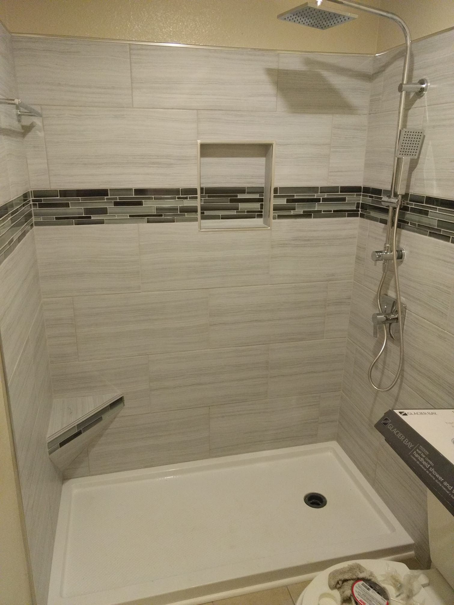 Newly tiled shower stall with white and gray tiles, showerhead, and a corner shelf.