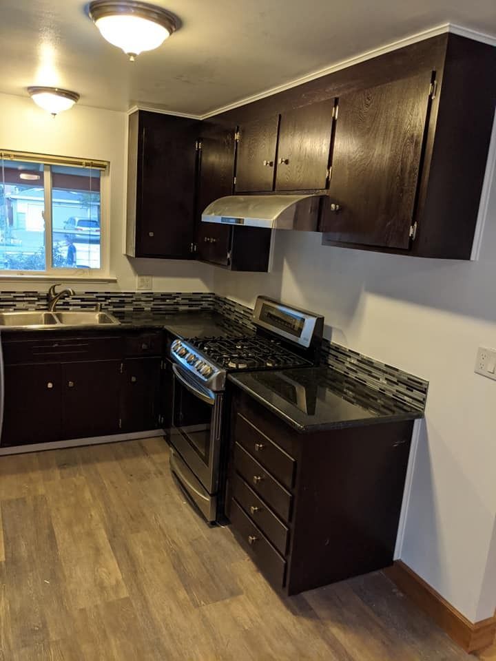 Dark kitchen with brown cabinets, stove, and sink. Light-colored flooring and small window.