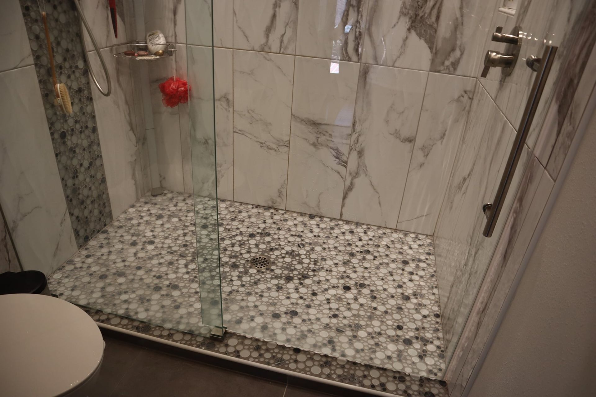 Shower with pebble floor, marble-like wall tiles, glass door. Soap and shower controls visible.