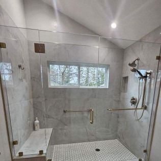 Glass-enclosed shower with gold fixtures, white tiled walls, window, built-in seat, and a pebble tile floor.