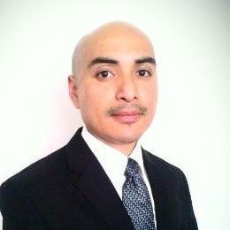 A bald man in a suit and tie is standing in front of a white wall.