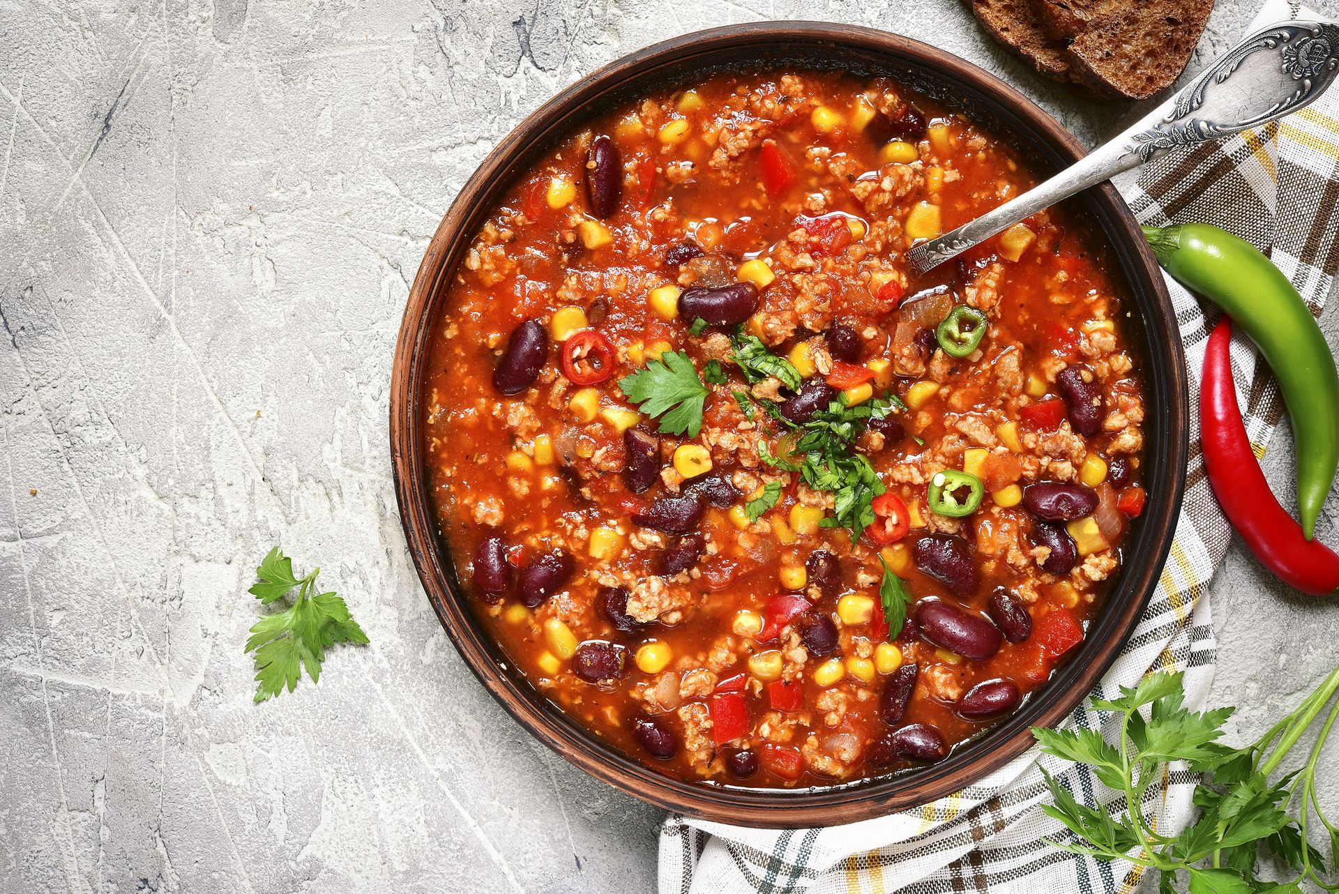A bowl of chili with red kidney beans, corn, and ground meat, garnished with herbs, next to green and red chili peppers.