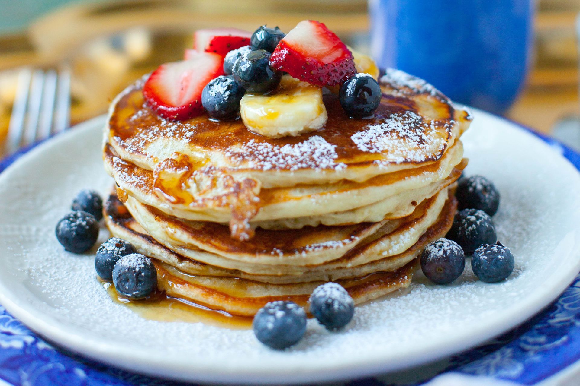 A stack of pancakes topped with strawberries, blueberries, banana slices, syrup, and powdered sugar on a blue-rimmed plate.