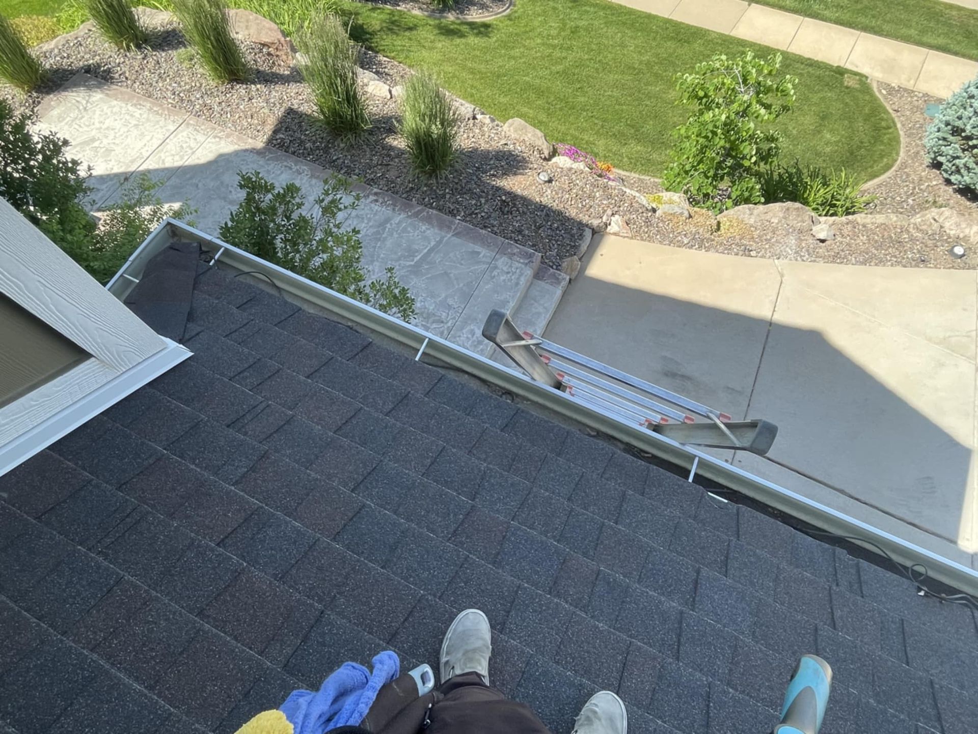 Person standing on a dark shingled roof, cleaning a gutter with a cleaning tool.