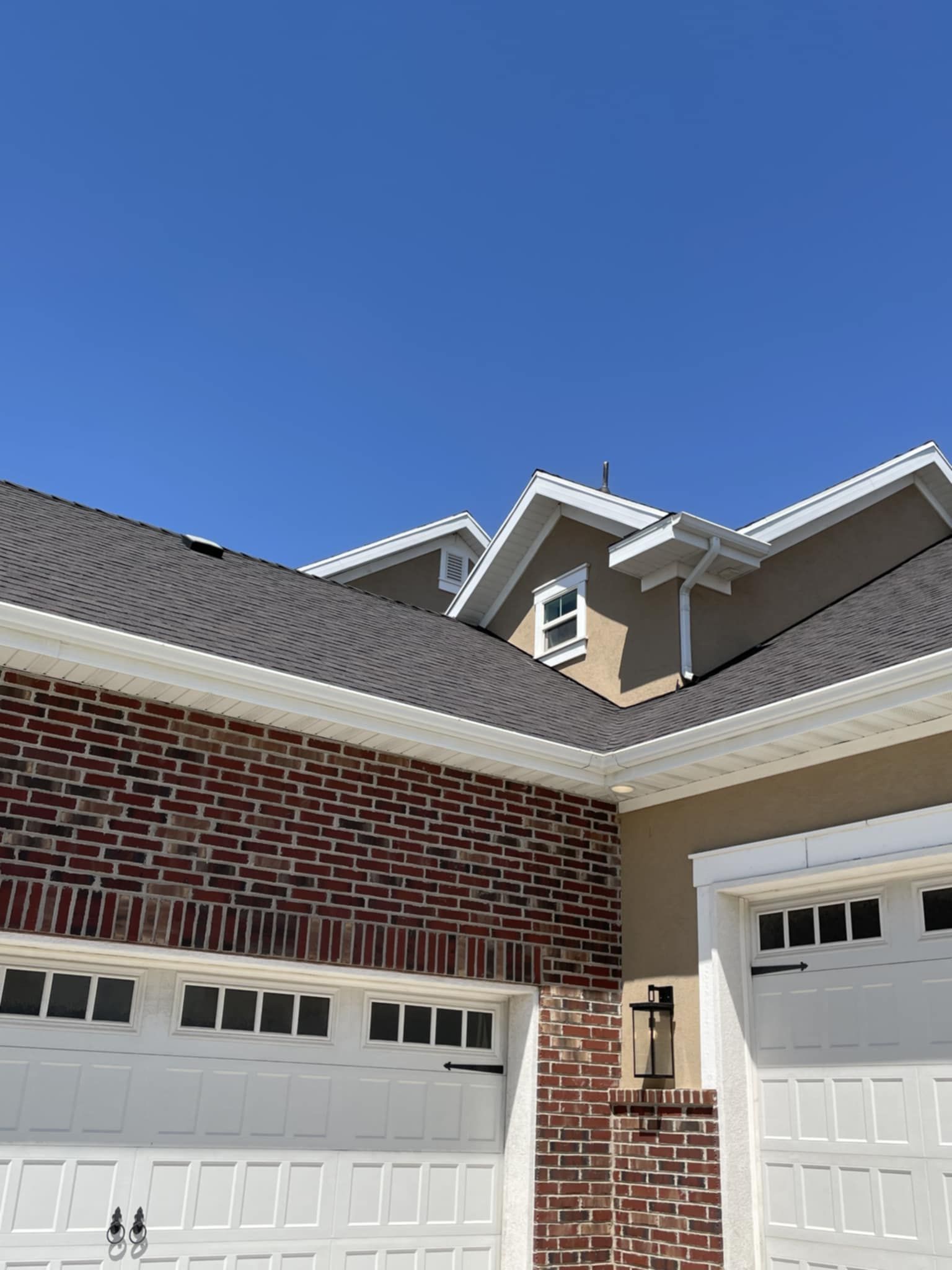 Brick and tan house exterior with white trim, white garage doors, and a dark roof against a clear blue sky.