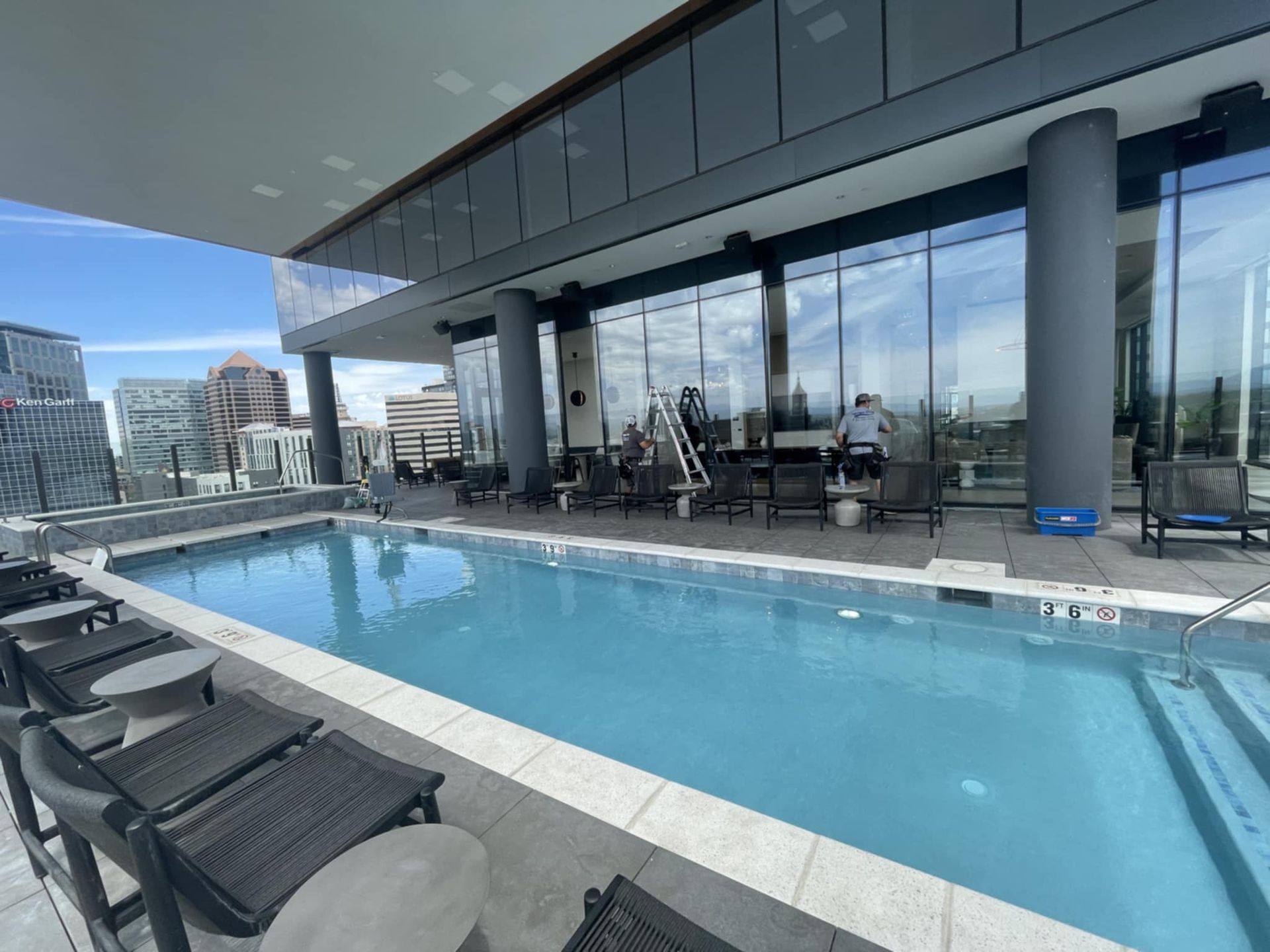 Pool on a rooftop patio with lounge chairs, overlooking a city. People are cleaning the large windows.