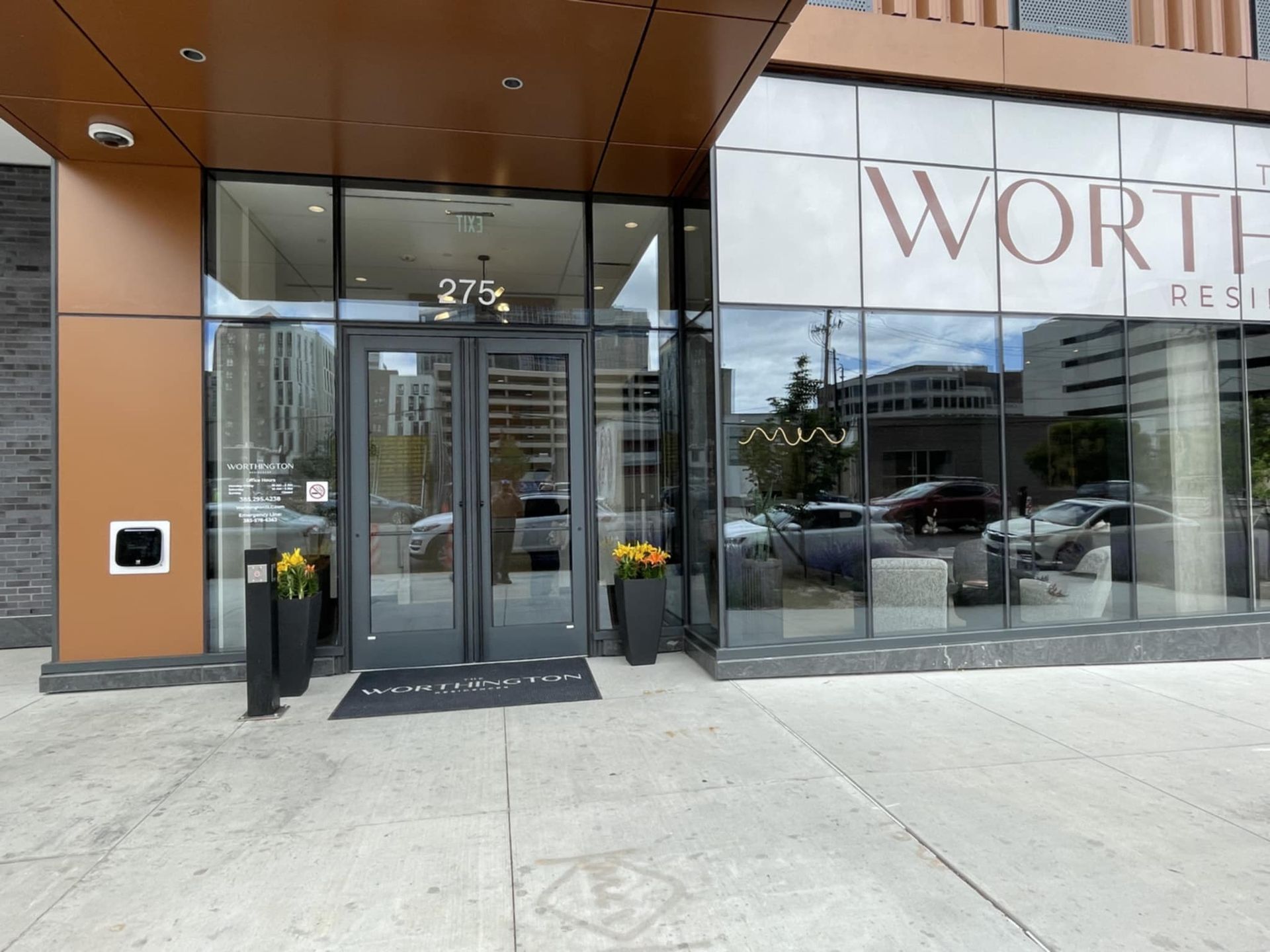 Entrance to The Worthington Residences, featuring glass doors, a doormat, and flower pots. Street view.