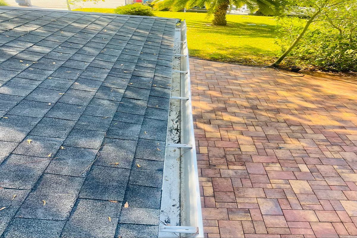 A gray asphalt shingle roof with a metal gutter alongside a red brick walkway.