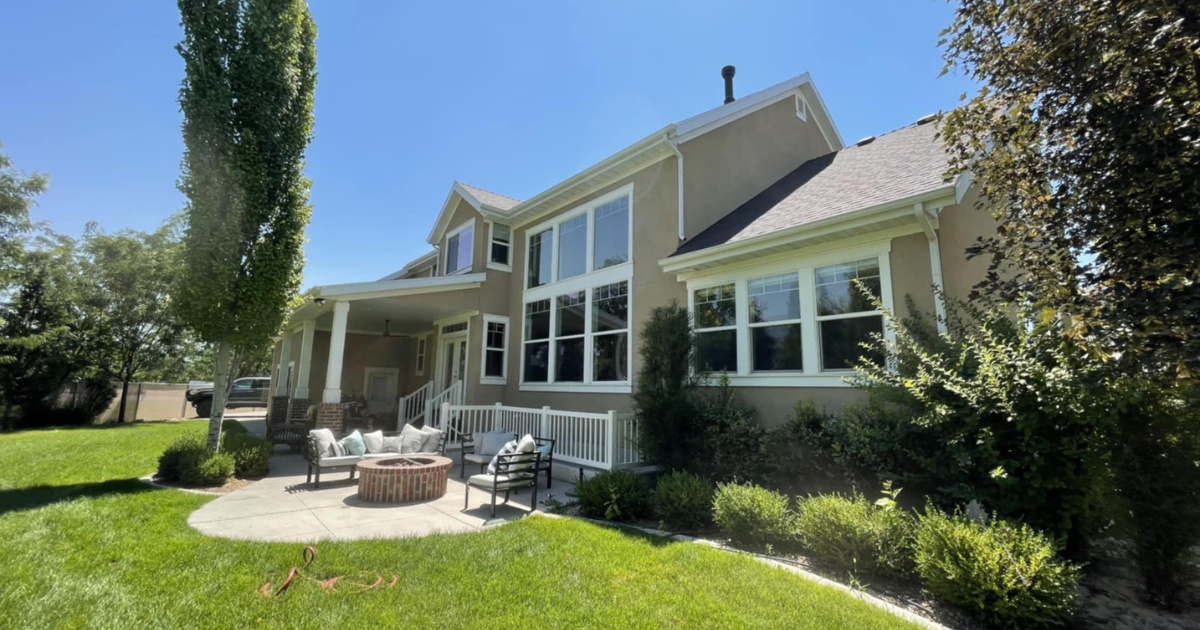 Beige house with patio, fire pit, and large windows, surrounded by green lawn and trees under a blue sky.