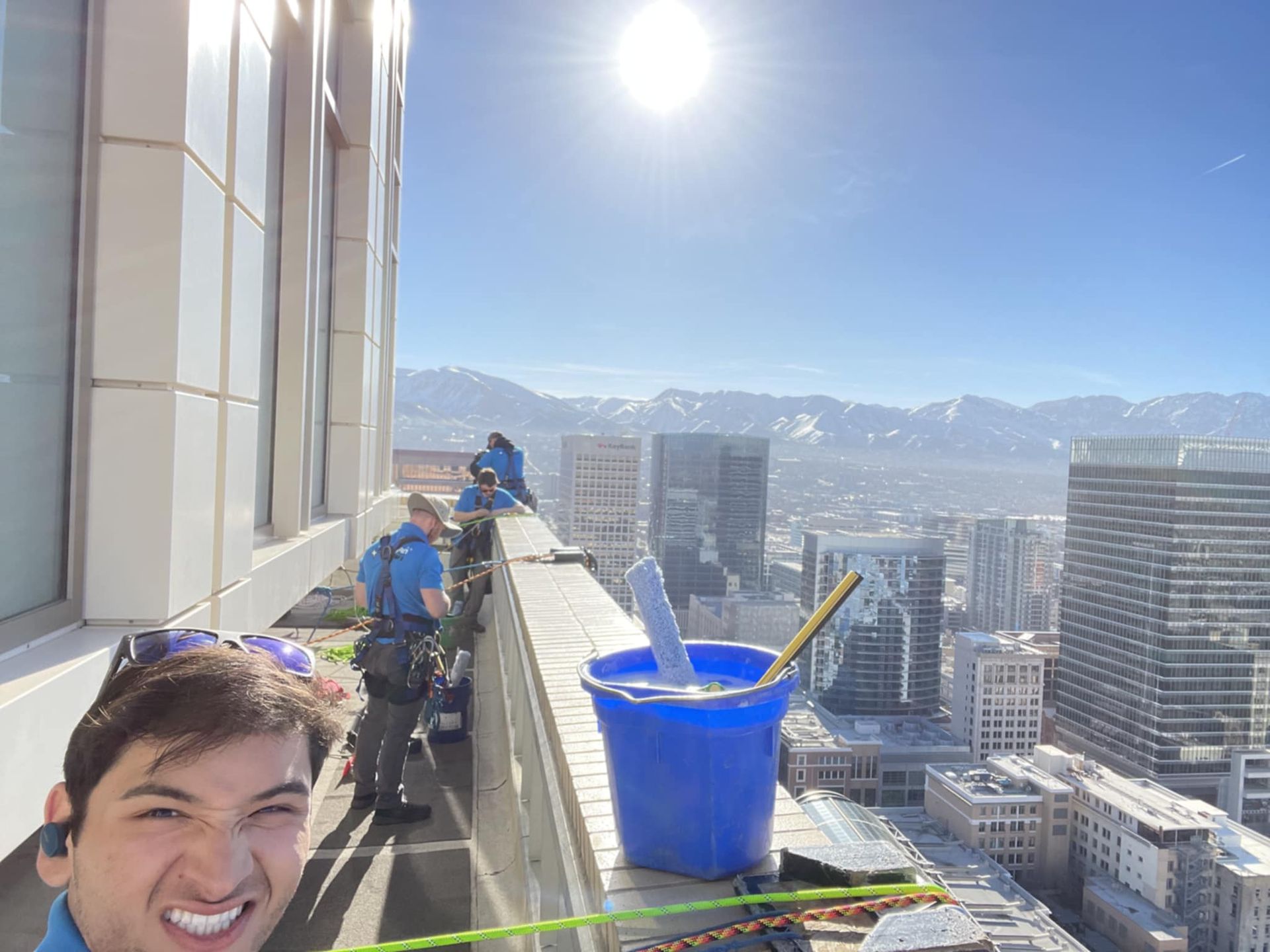 Man taking selfie on a high-rise building ledge with window washers, blue sky, and city view.