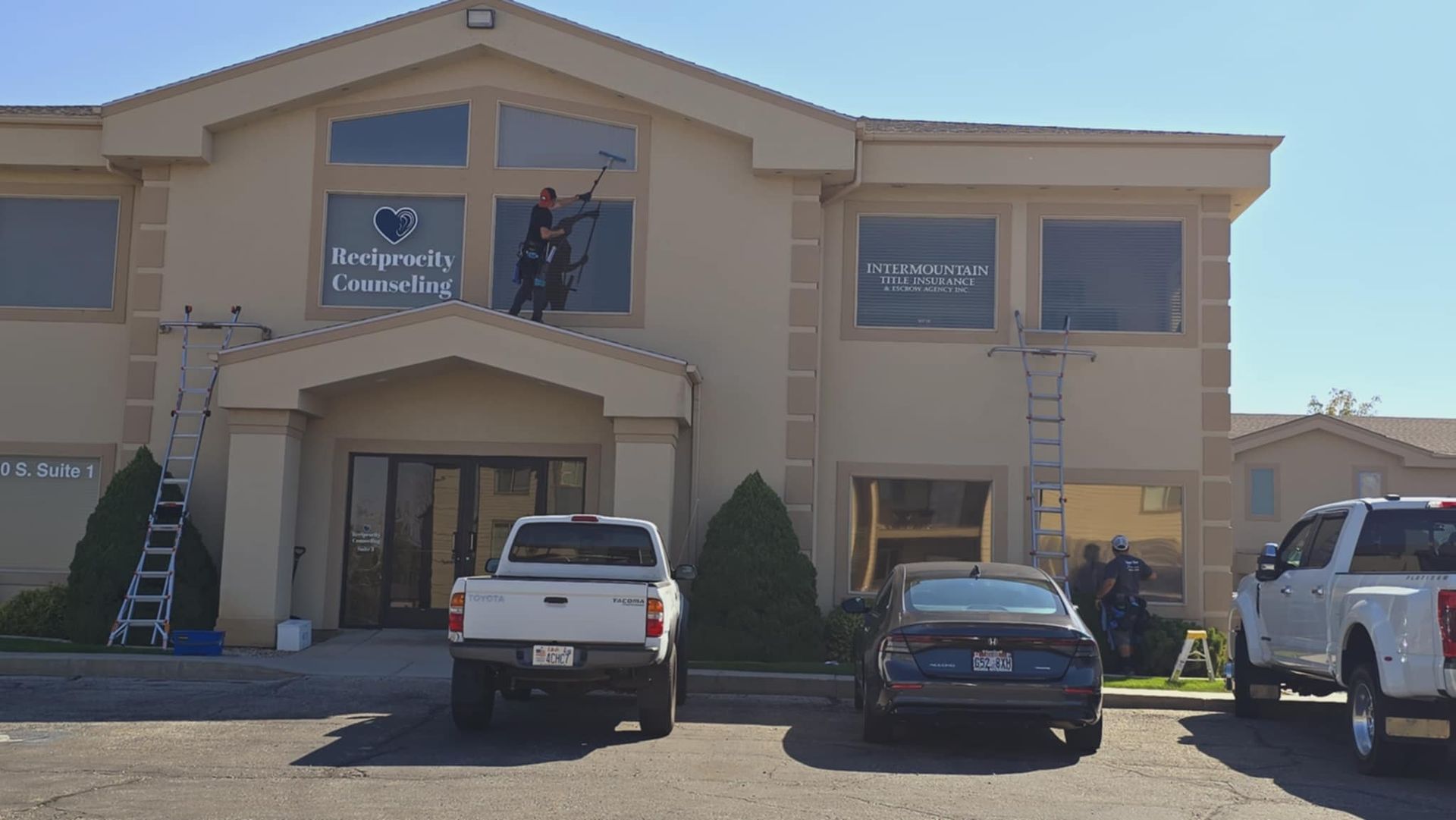 Window washers cleaning the windows of a two-story tan building. Trucks and cars parked in front.
