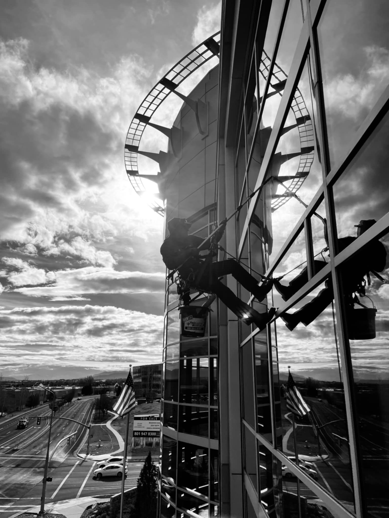Window washers suspended on a high-rise building, silhouetted against a bright, cloudy sky over a city.