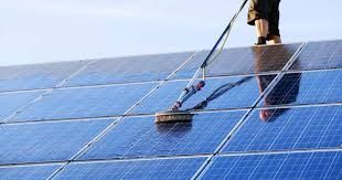 Person cleaning solar panels on a rooftop with a long-handled brush and water spray, blue panels and clear sky.