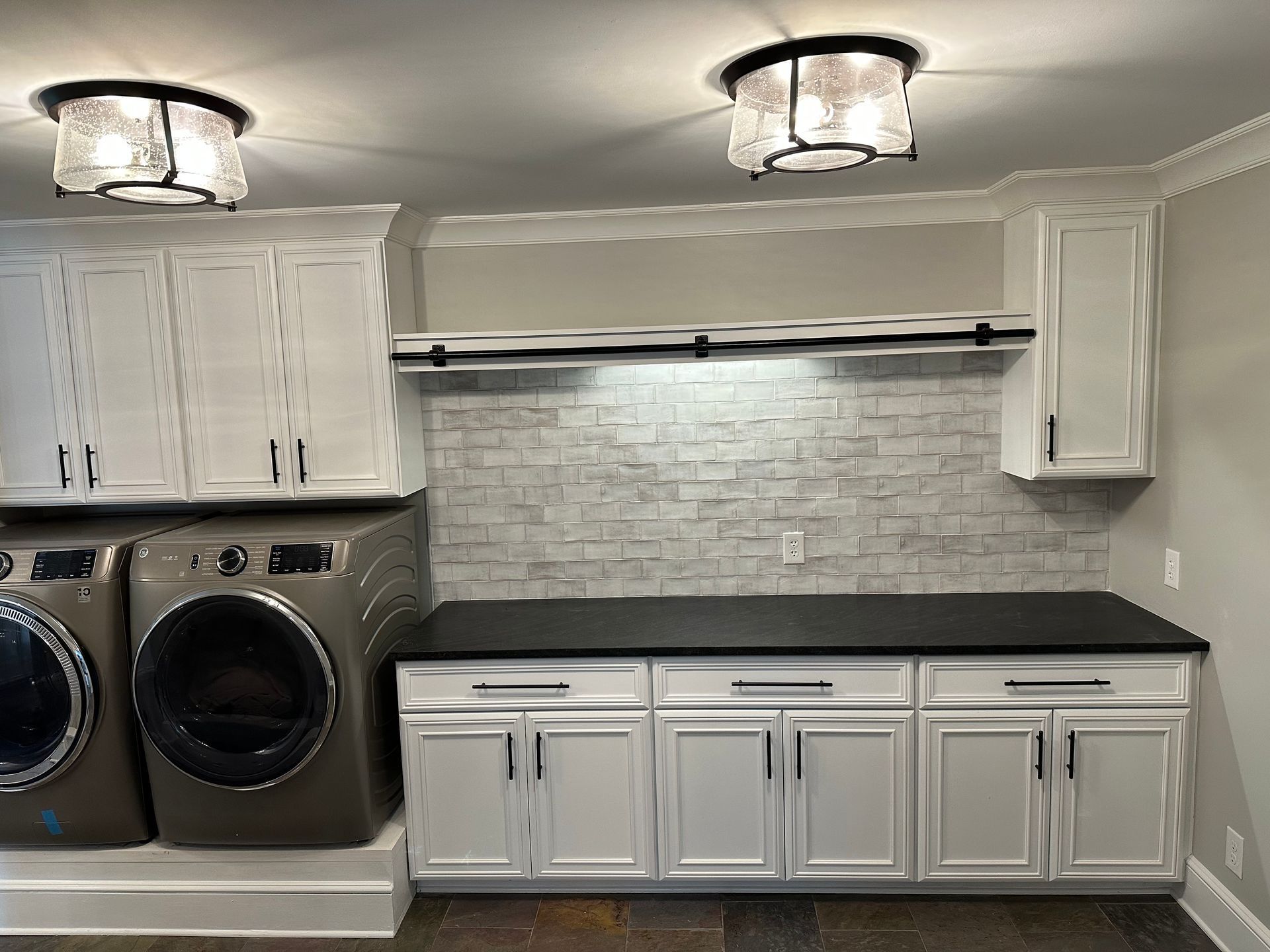 Laundry room with white cabinets, dark countertop, and silver appliances. Gray brick backsplash.