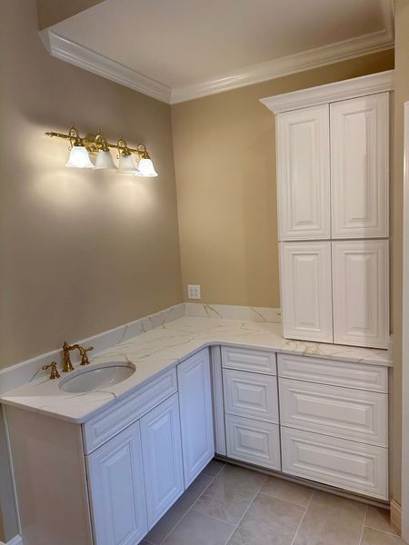 White bathroom vanity with marble countertop, sink, and upper cabinets, gold fixtures, and wall-mounted lights.
