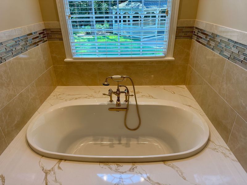 Oval bathtub in a beige tiled bathroom, with a window and gold faucet.