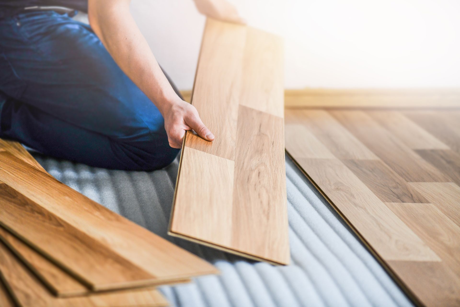 Person installing wood flooring, kneeling, holding plank, light brown planks.