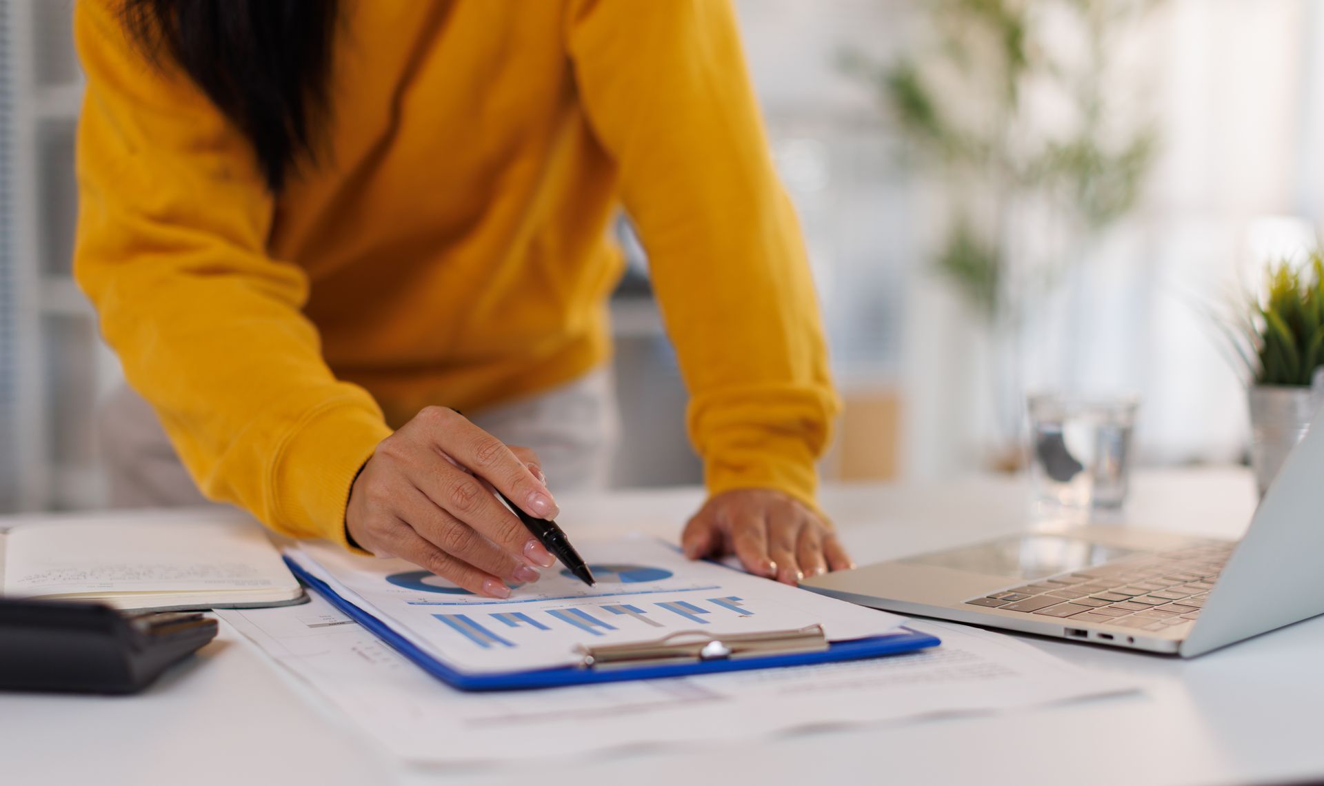 Person in a mustard sweater points to charts on a clipboard beside a laptop at a desk Person in a mustard sweater points to charts on a clipboard beside a laptop at a desk