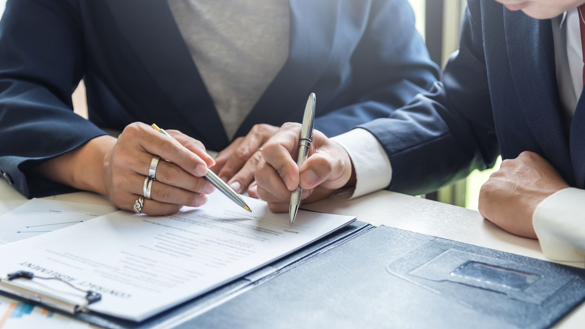 Two people signing documents at a table with a laptop nearby Two people signing documents at a table with a laptop nearby
