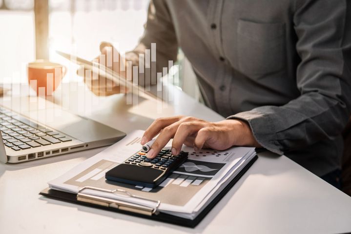 Person using a calculator on a clipboard beside a laptop, with financial charts in the background Person using a calculator on a clipboard beside a laptop, with financial charts in the background