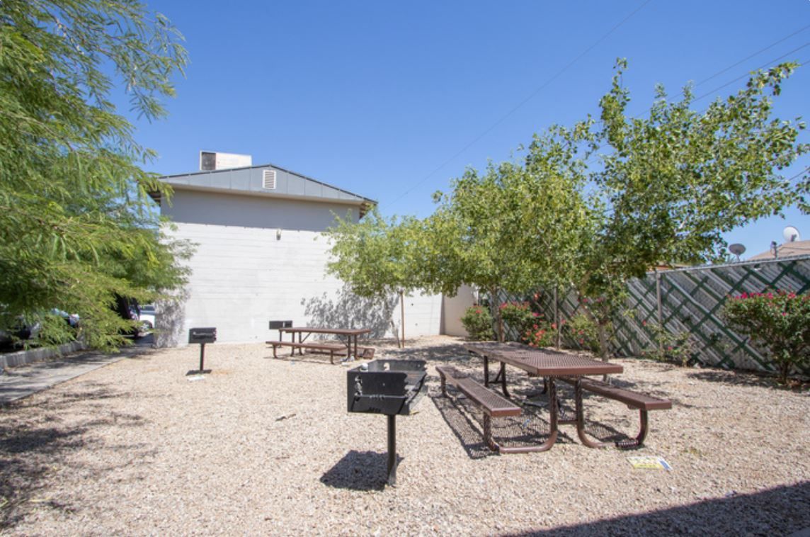 Gravel patio with two picnic tables and a grill under trees, beside a white building on a sunny day.