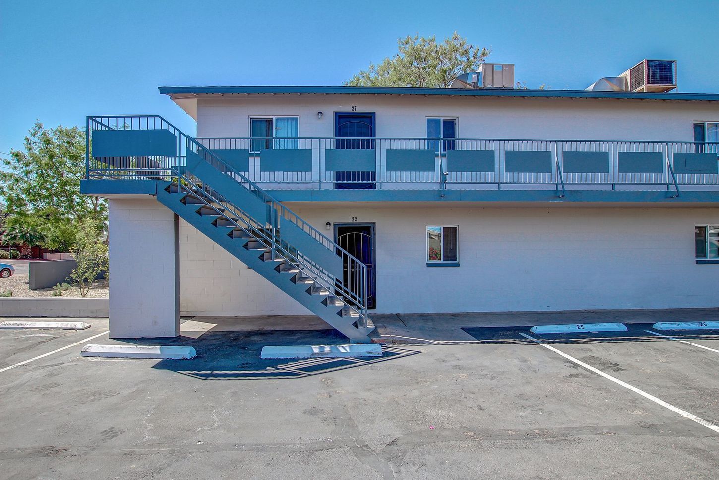 Two-story apartment building with exterior stairs and parking spots, blue railing, cream walls, clear sky.