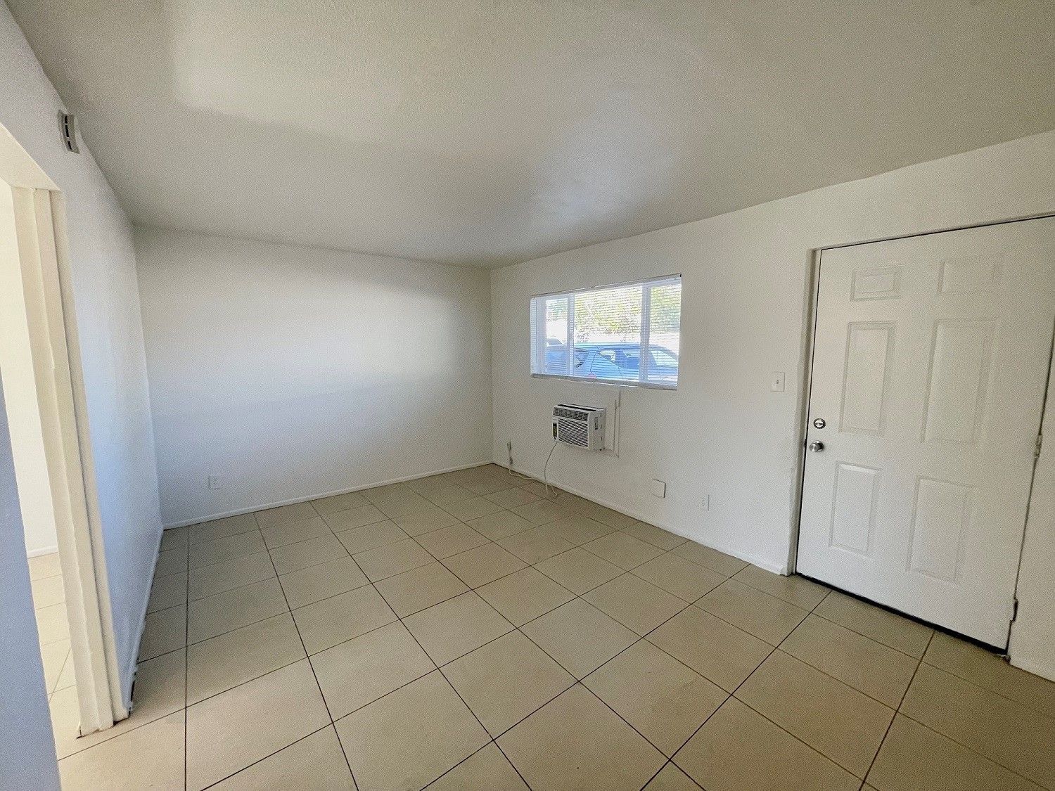Empty room with white walls, tiled floor, door, window with an AC unit, and an open doorway.