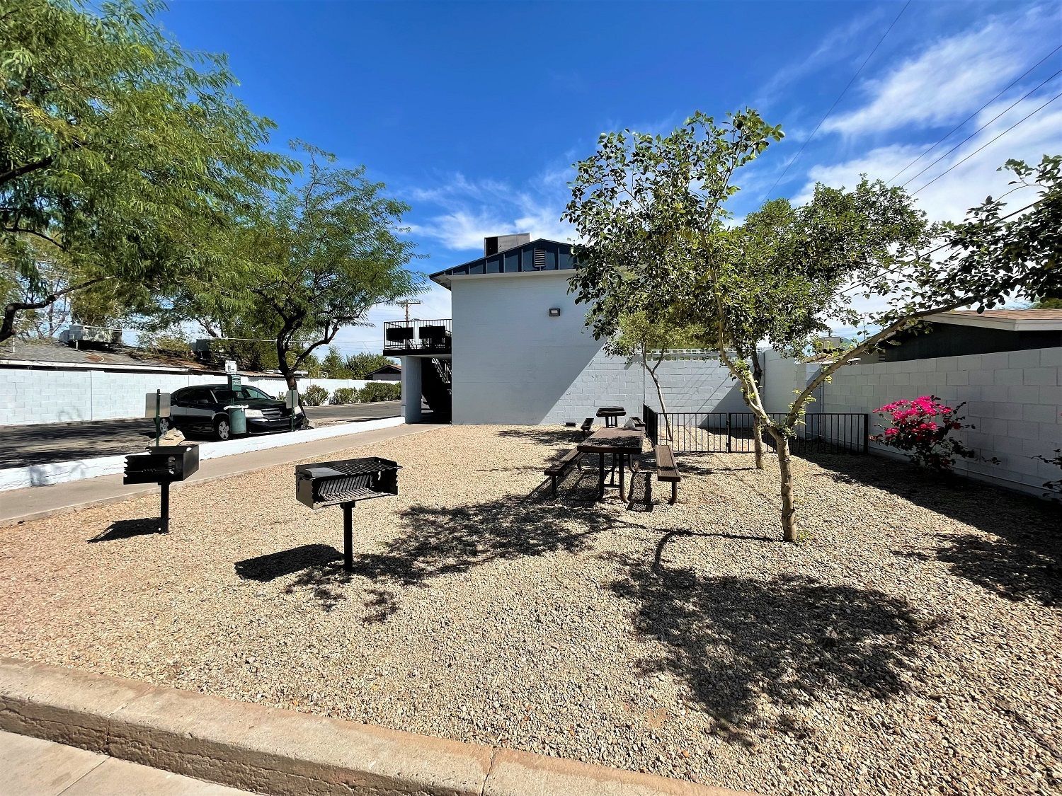 A small outdoor area with two grills, picnic tables, and trees under a blue sky.
