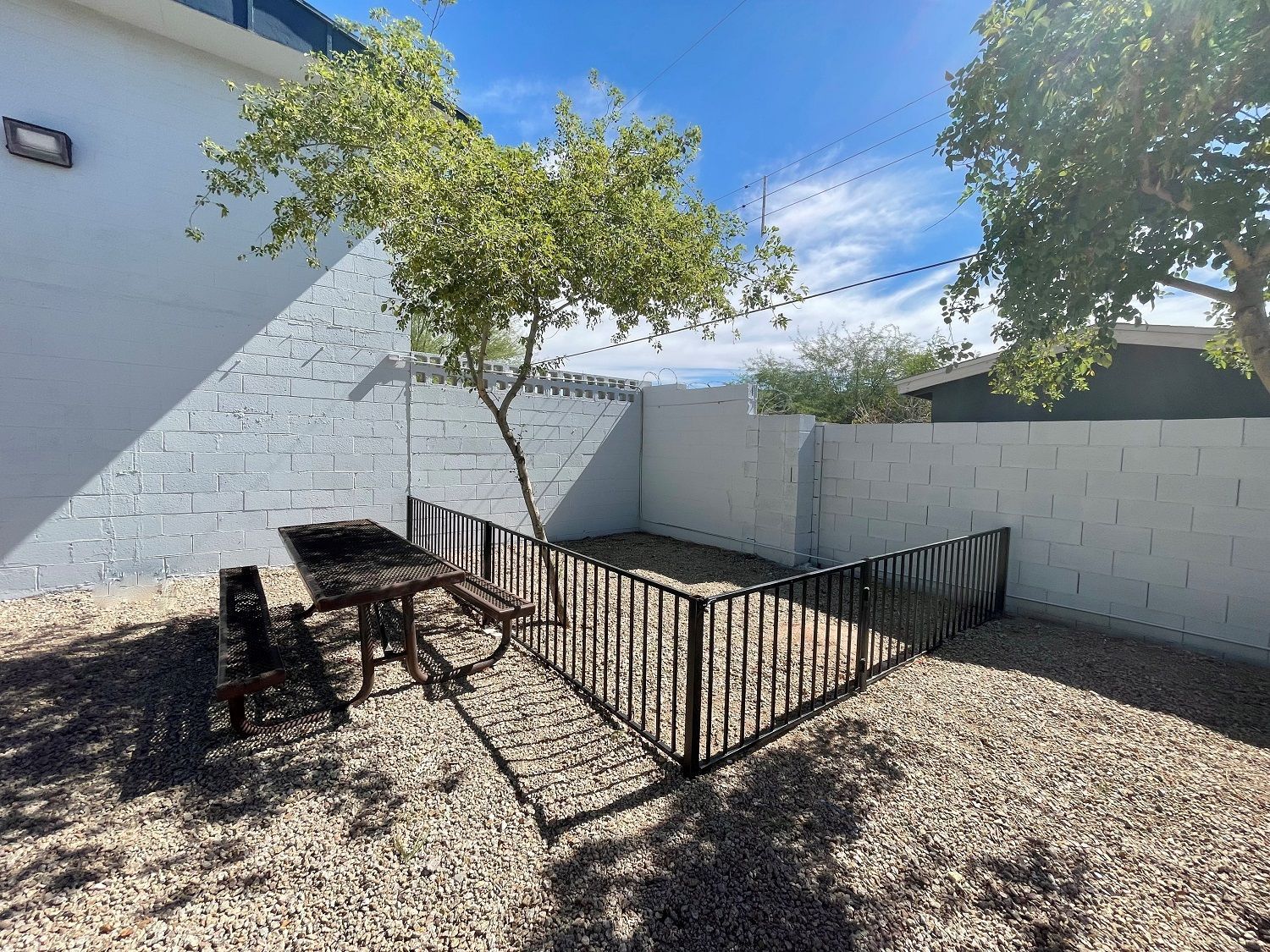 Picnic table in a gravel yard, enclosed by black fence, with trees and a white wall.