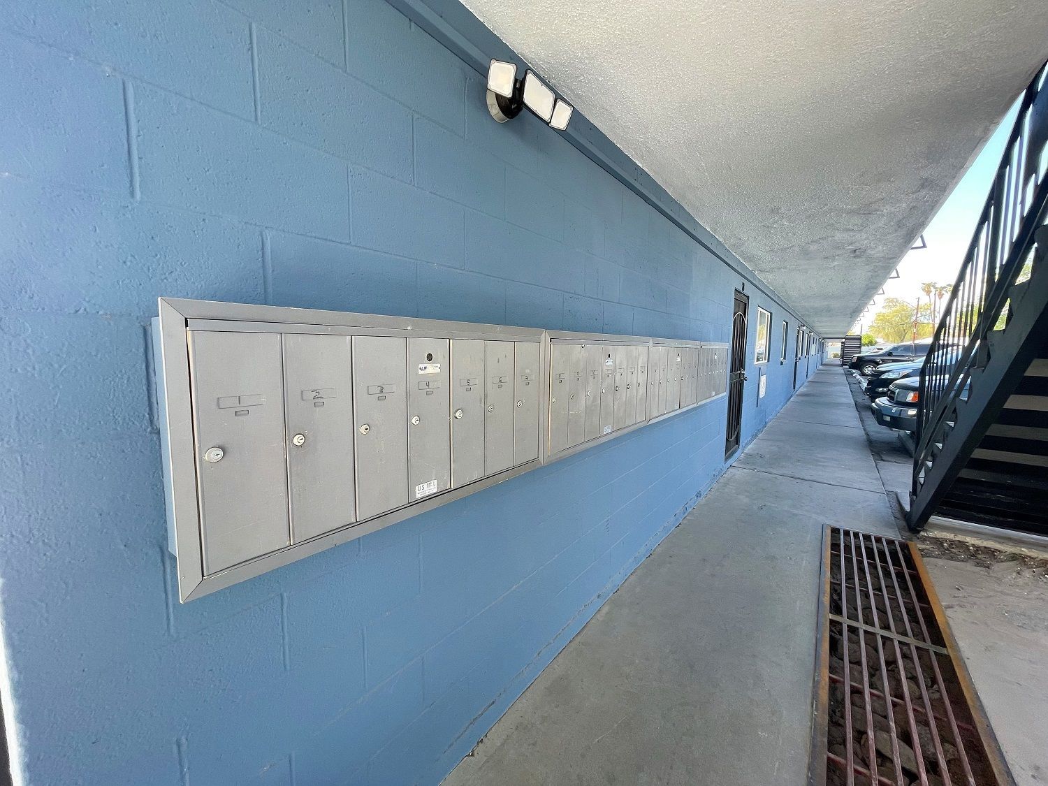Mailboxes built into a blue wall, located in a walkway with stairs and cars visible in the background.