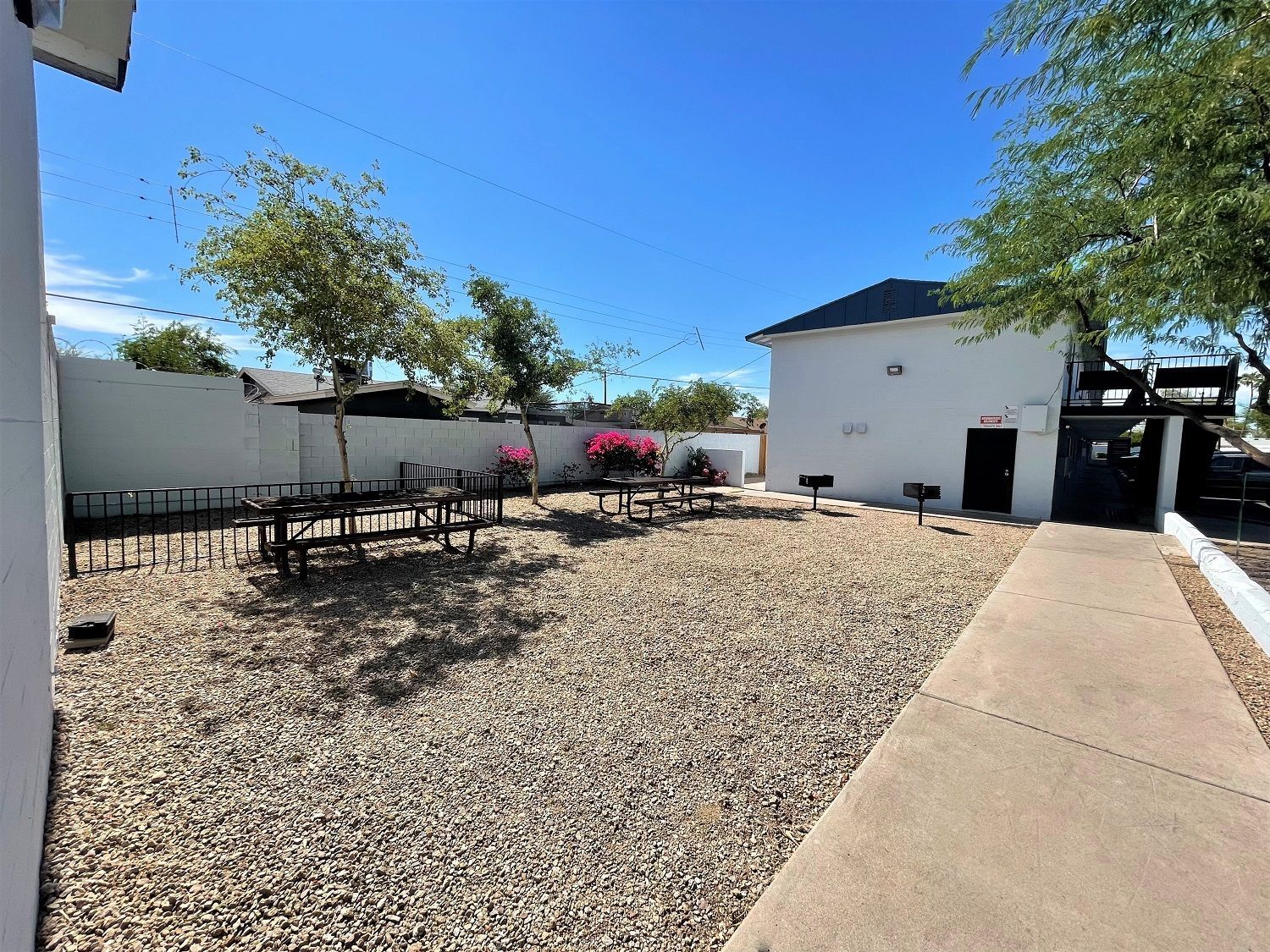 Outdoor area with picnic tables, small trees, gravel ground, white building, and blue sky.