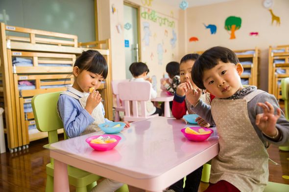 Children at a pink table eating fruit in a classroom. One boy gives the OK sign.