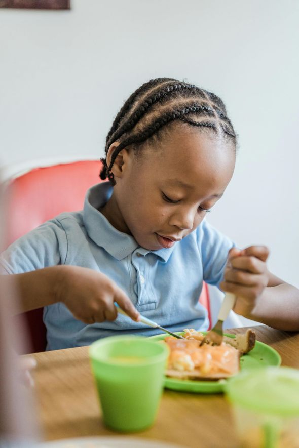 Young child with braids eats at a table, using a fork and knife.