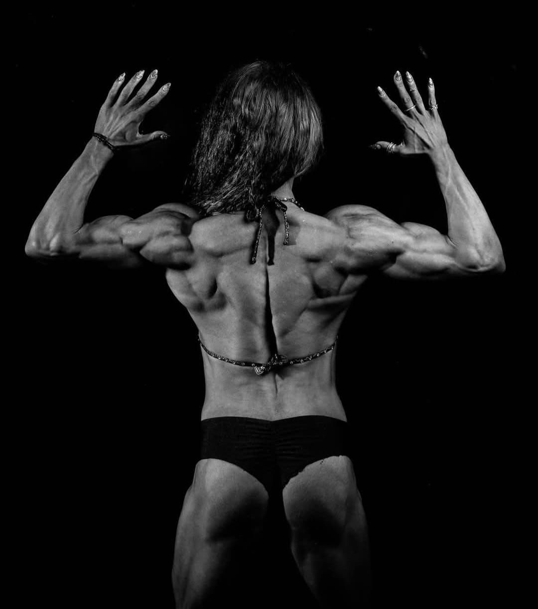Female bodybuilder flexing her back muscles in black and white, posing against a dark background.