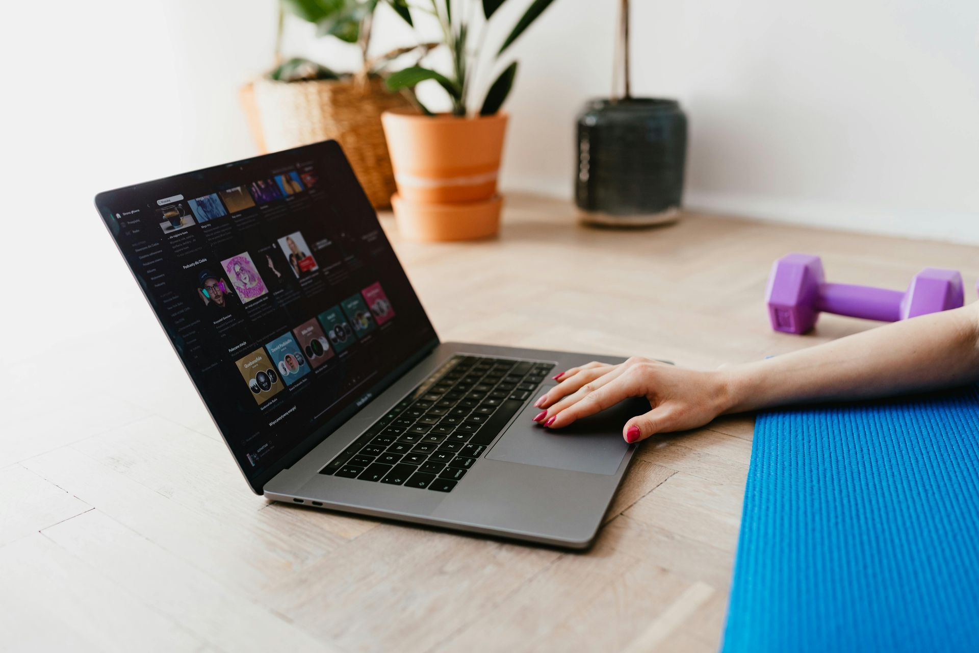 Person using laptop on floor, a yoga mat and weights nearby, with plants in background.