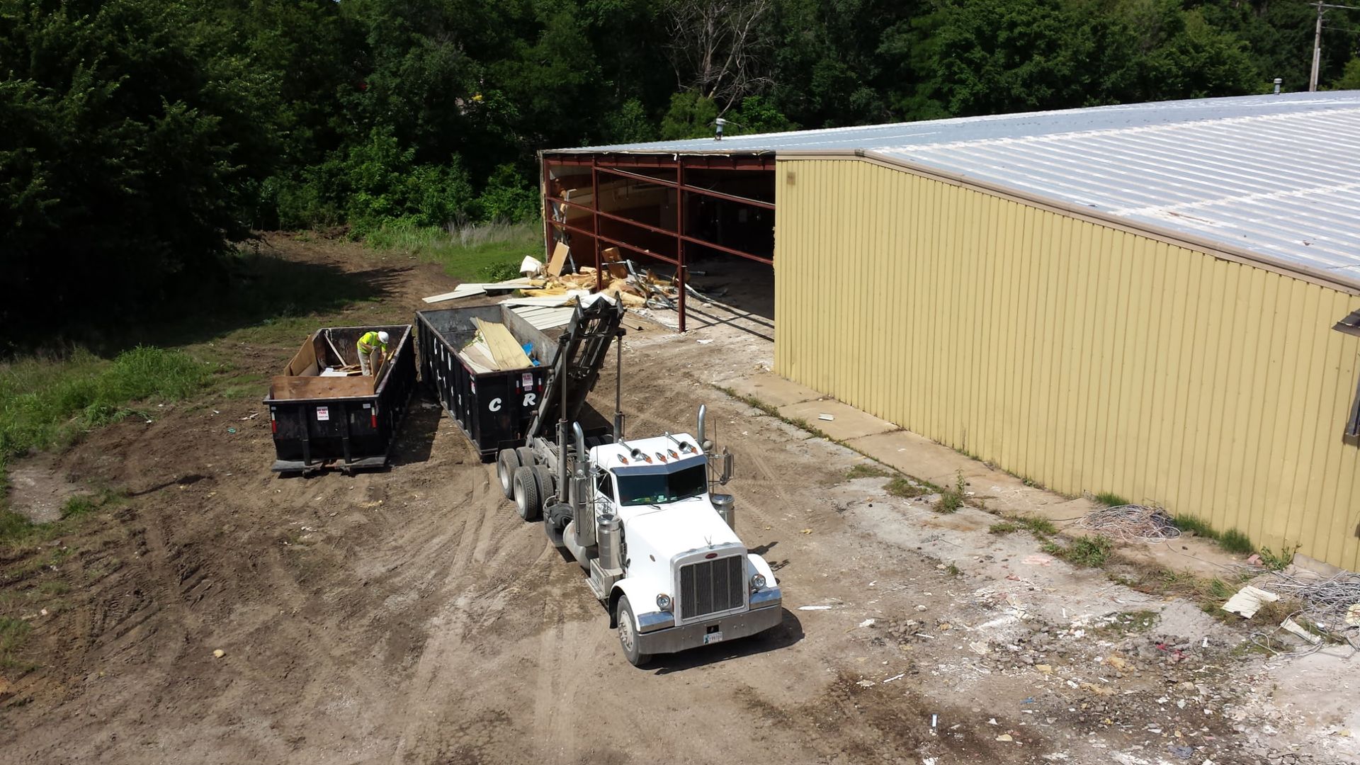 An aerial view shows a white truck loading debris into a dumpster beside a large yellow industrial warehouse.