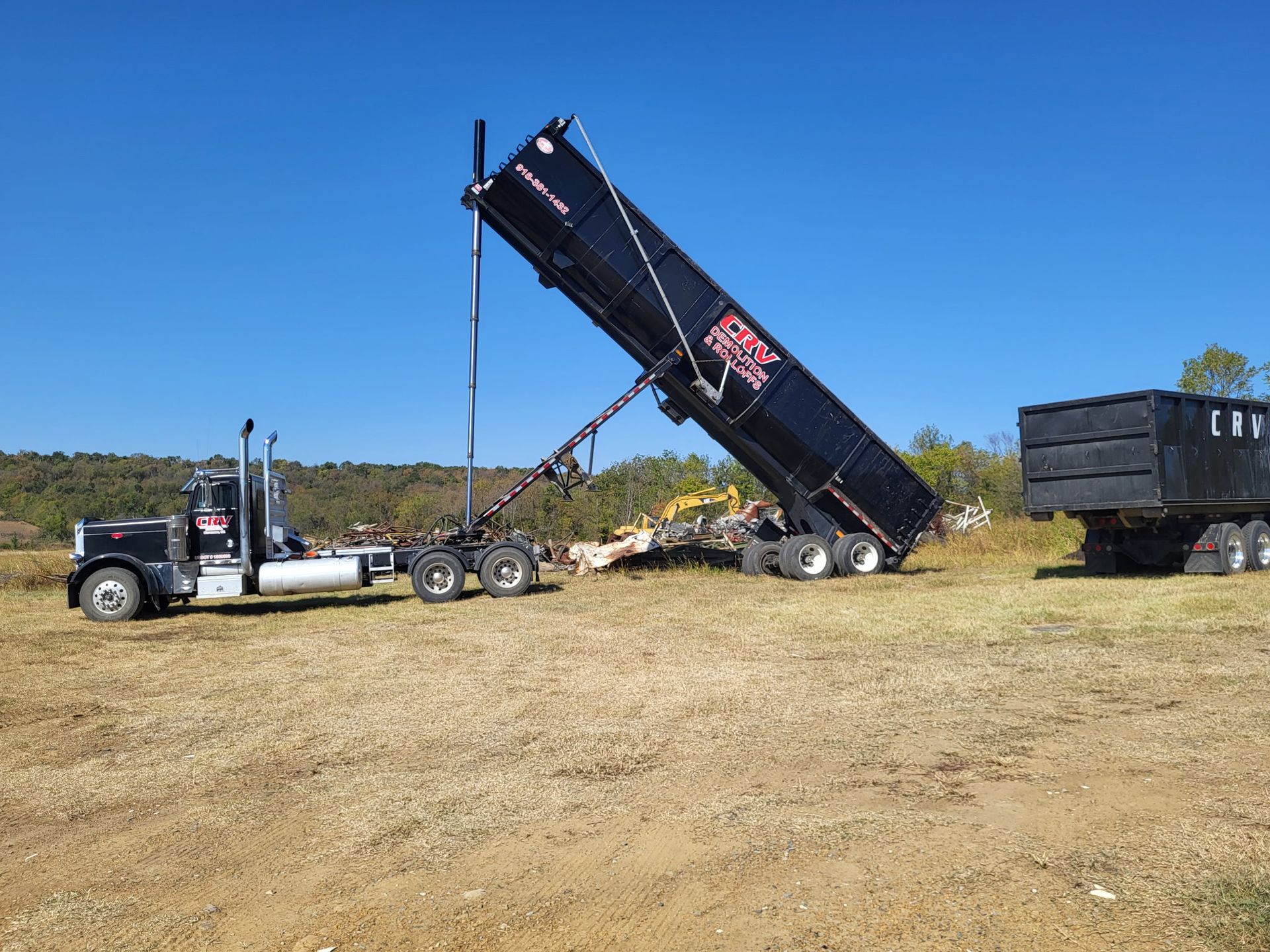 A black dump trailer raised to unload onto a field, with a semi-truck connected and a second trailer parked nearby.