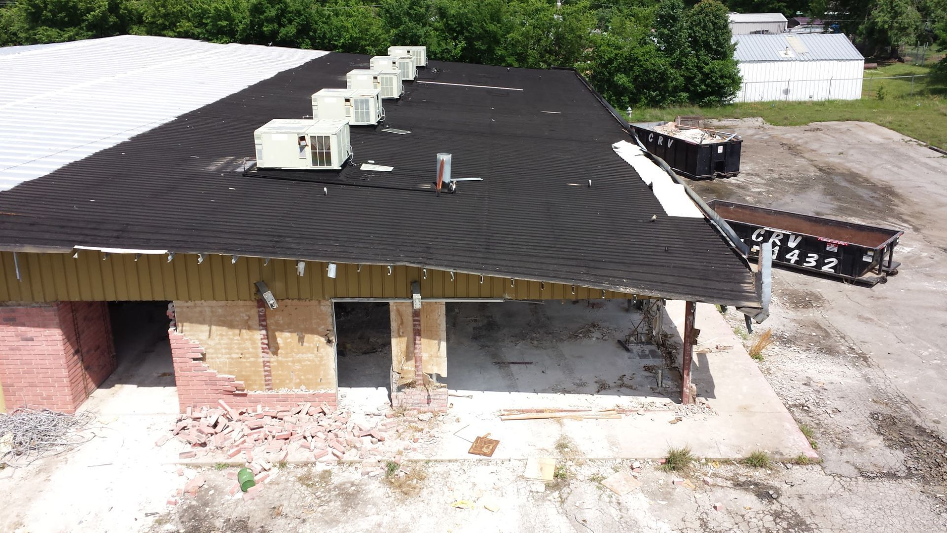 An aerial view shows a commercial building under renovation with exposed wood framing, a black roof, and dumpsters nearby.