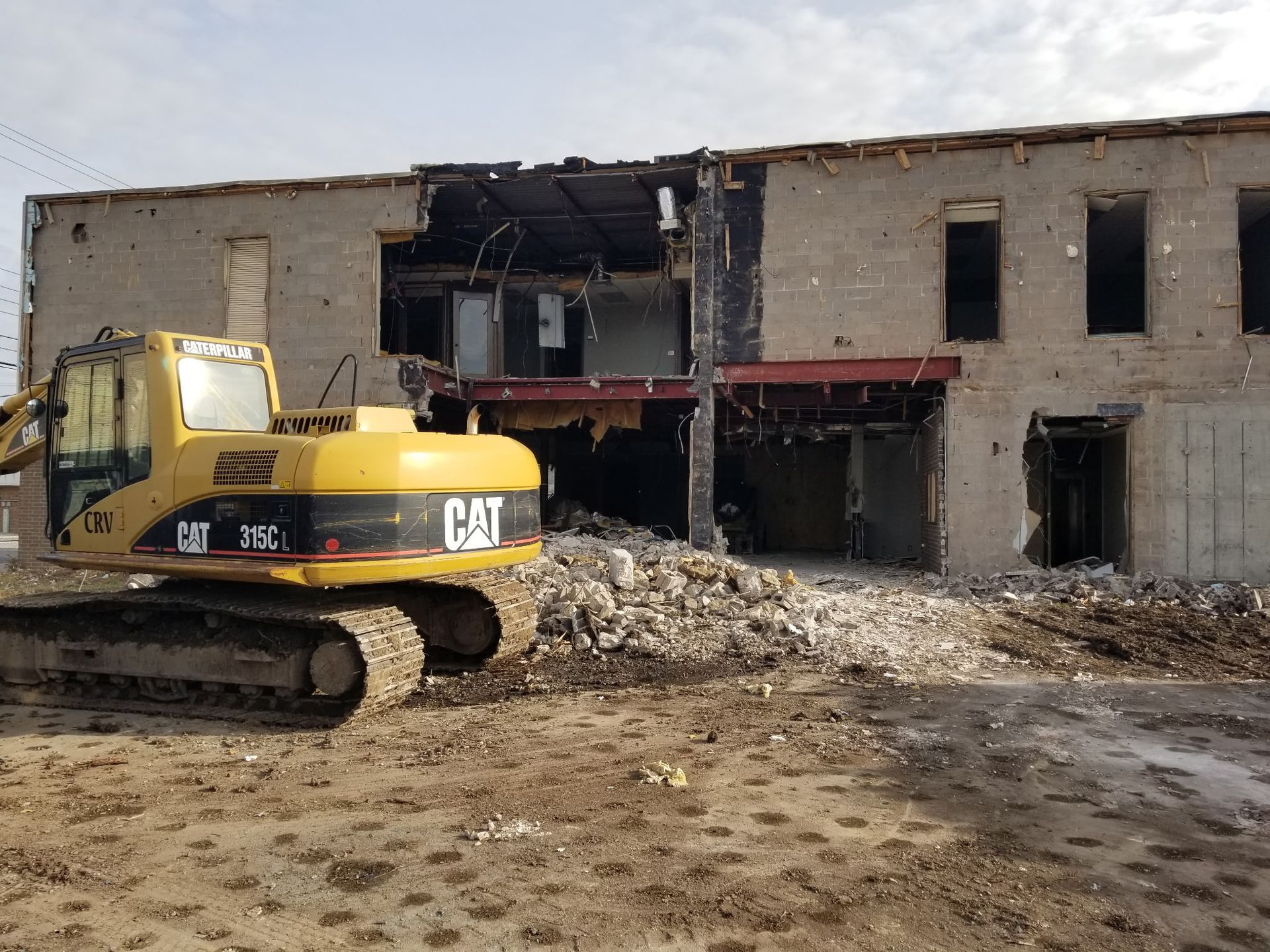 A yellow CAT excavator sits in front of a multi-story building undergoing demolition with rubble piled in the entryway.