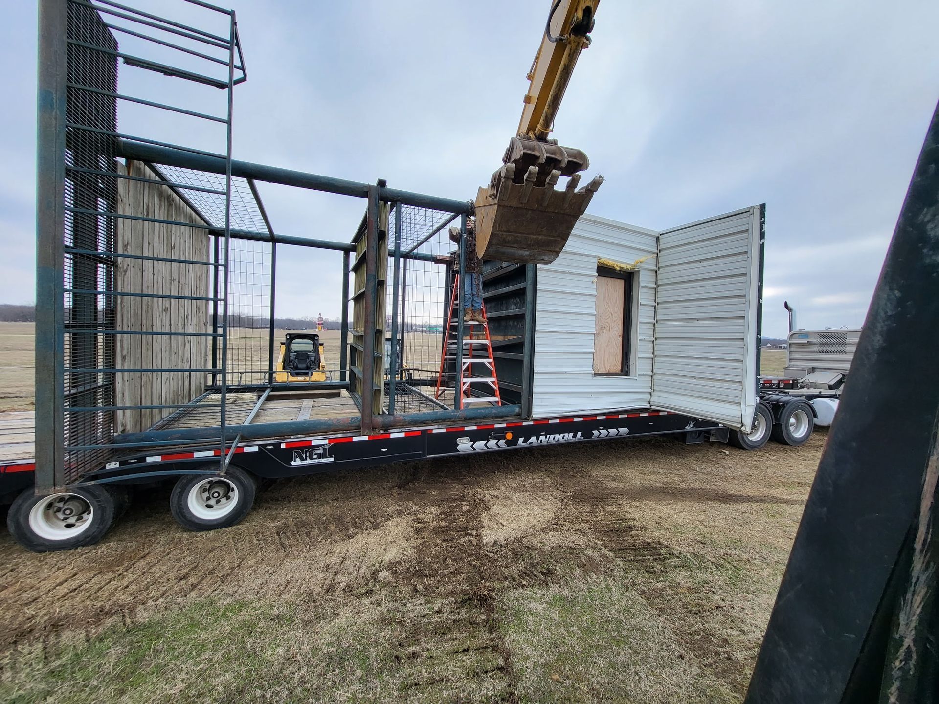 An excavator lifts a wall panel onto a steel structural frame mounted on a flatbed trailer in a rural field.