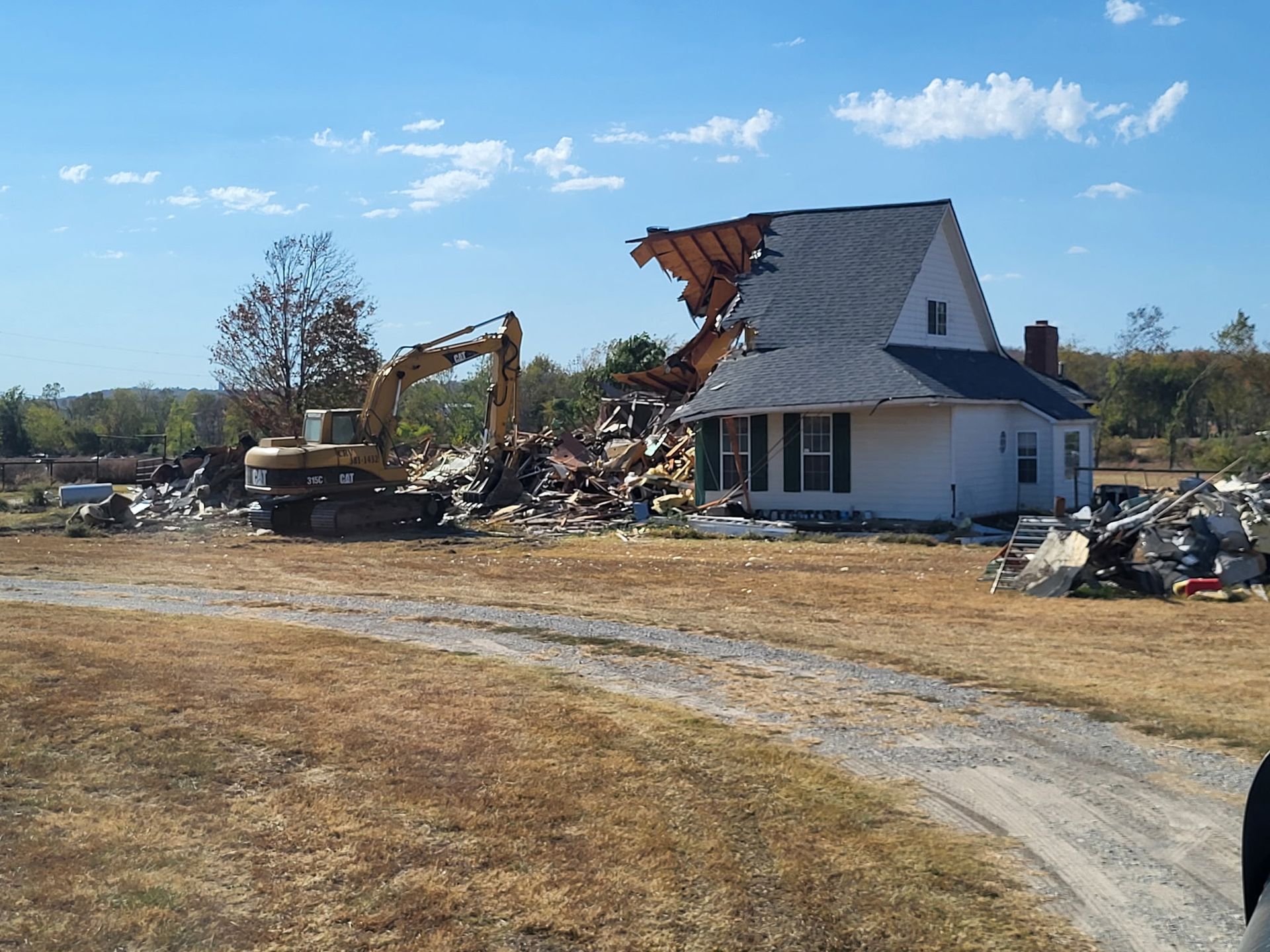 An excavator tears down a house on a clear day, with rubble scattered in a grassy field.