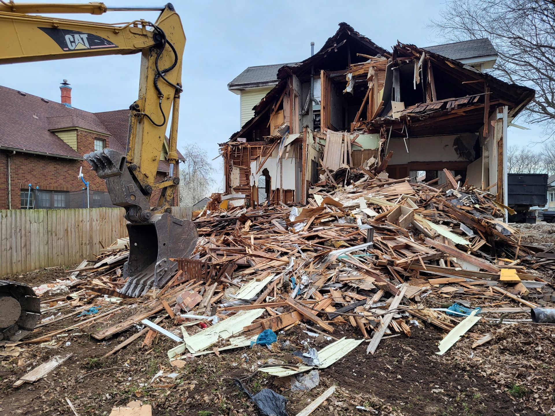 A yellow CAT excavator dismantling a house, with debris scattered in the foreground against a cloudy sky.