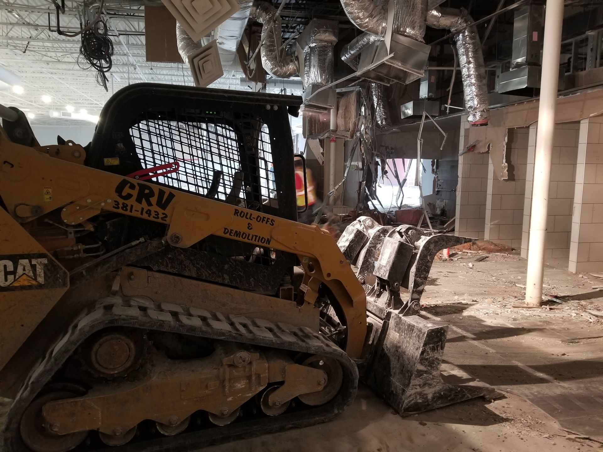 A yellow CAT skid-steer loader sits inside a building undergoing demolition with exposed ceiling ducts and debris.