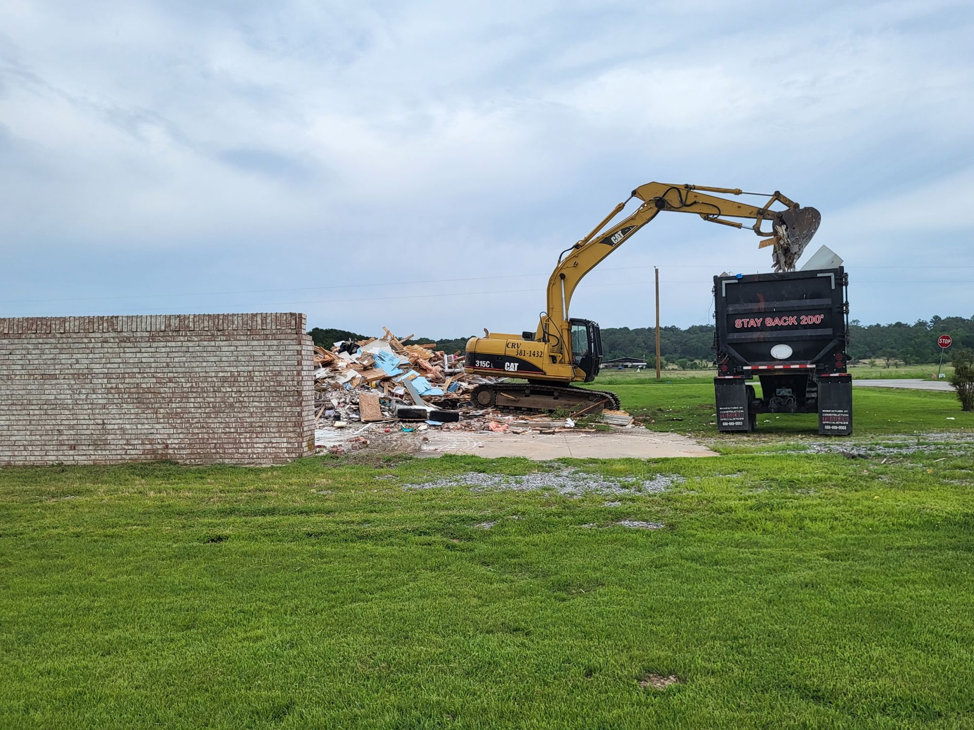 A yellow excavator loads debris into a black dump truck next to a brick wall in a grassy field under a cloudy sky.