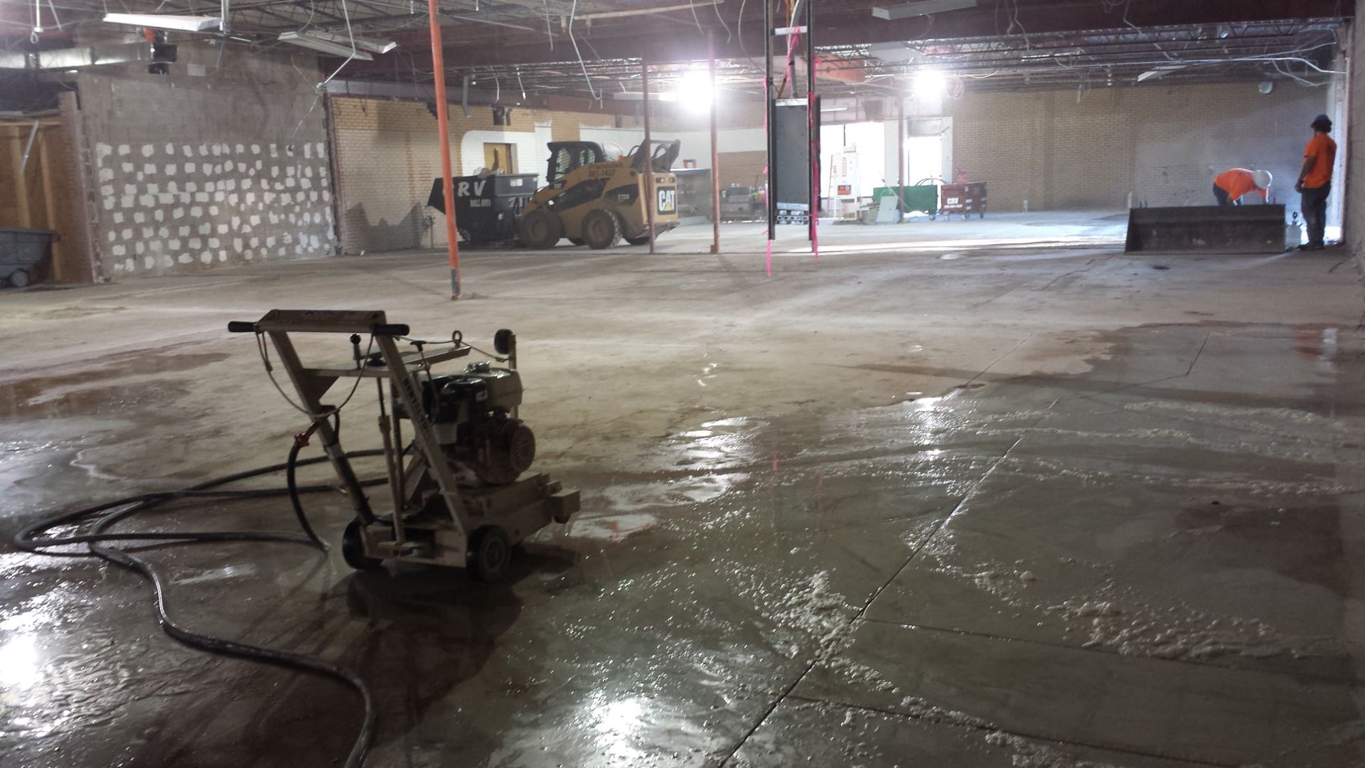 A construction crew works in a large, unfinished interior space with a floor polisher in the foreground.